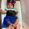 baby, infant, headband, blue_skirt, black_top, changing_pad, blanket, clothing, cute, child, person, indoor, soft_texture, pink_clothing, sequin_bow, reaching_hand, tongue_out, foot, lying_down, wooden_frame