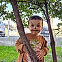 child, face_paint, tree, brown_clothing, grass, outdoor, fence, car, young_child, playful, smile, nature, portrait, casual_wear, daylight, curious, person, leaves, standing, expression