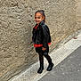 black_boots, black_skirt, casual, child, confident, fashion, footwear, hair_tied_back, happy, leather_jacket, outdoor, person, posing, red_top, sidewalk, smiling, street, urban, wall, young