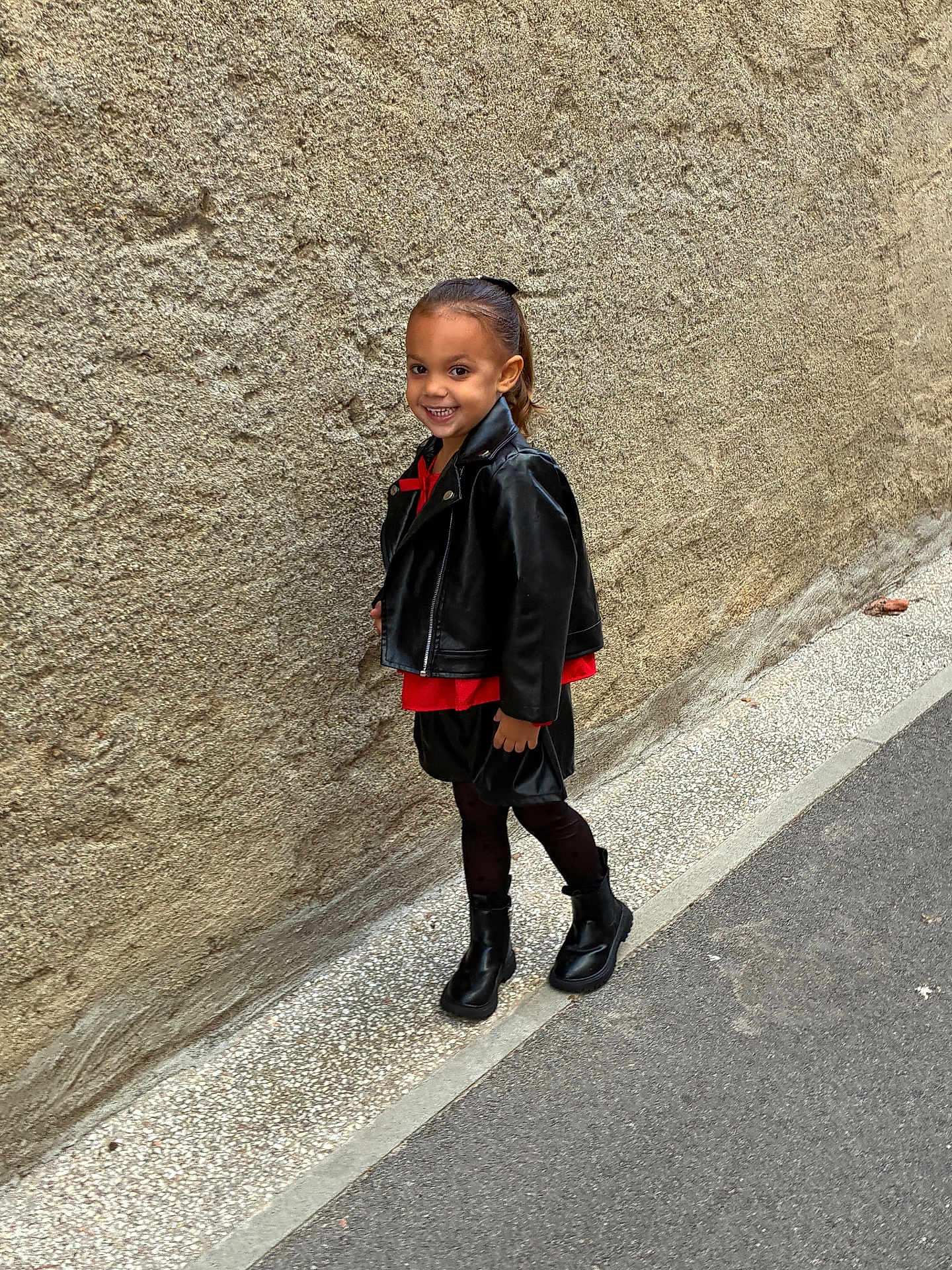 Tya a rejoint le concours — aidez-le/la à gagner de superbes lots ! child, smiling, leather_jacket, red_top, black_skirt, black_boots, street, sidewalk, wall, urban, fashion, posing, happy, person, young, outdoor, confident, casual, footwear, hair_tied_back