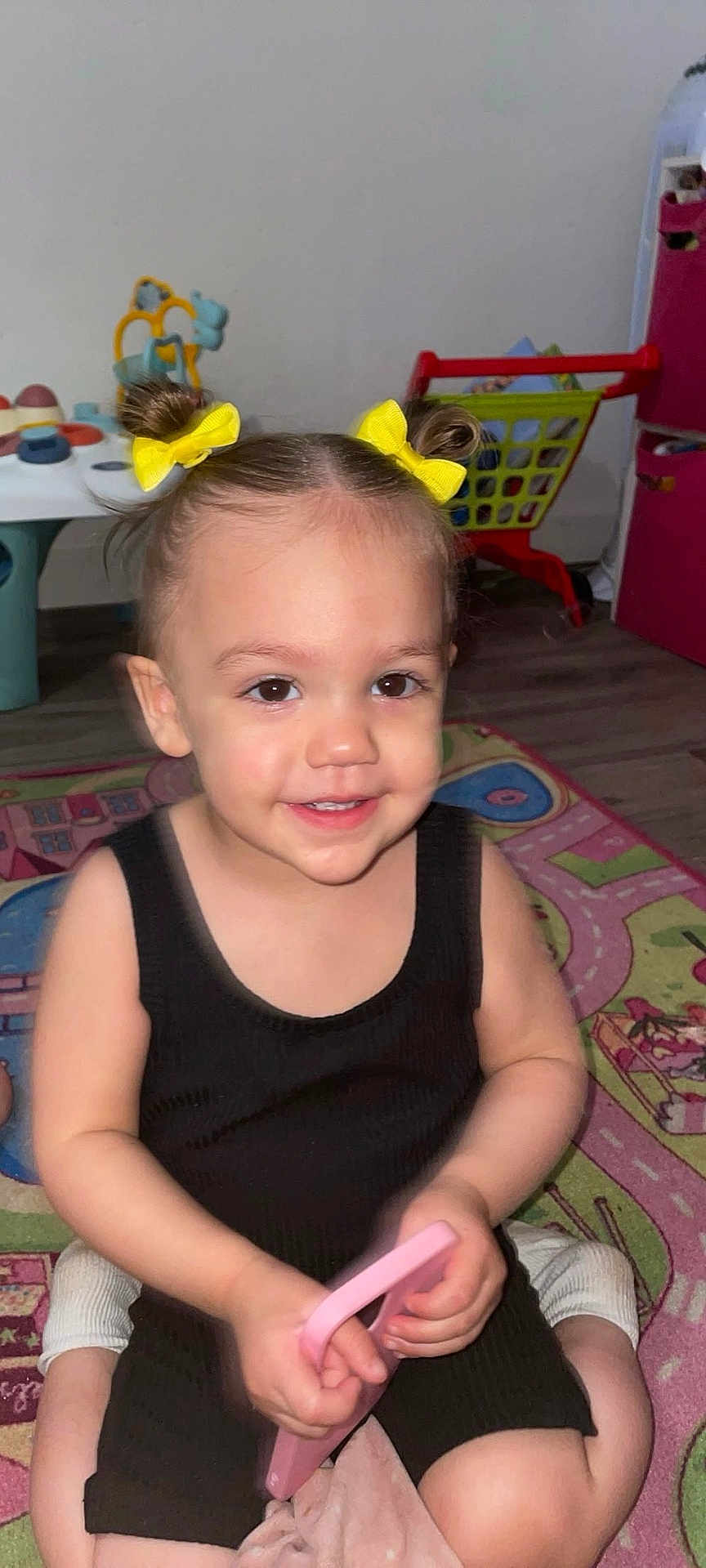 Ava participe au concours pour gagner de l'argent avec cette photo : toddler, child, yellow_bows, black_outfit, pink_toy, play_mat, indoor, smiling, happy, toys, shopping_cart, floor, hair_buns, person, cute, young_child, playtime, colorful, room, carpet