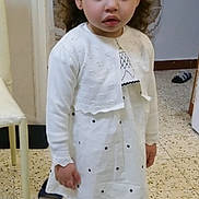 Aicha participe au concours pour gagner de l'argent avec cette photo : child, girl, white_dress, curly_hair, flower_hair_accessory, cardigan, tights, shoes, indoor, tile_floor, patterned_wall, chair, slipper, standing, curious_expression, person, young_child, home_interior, footwear, casual