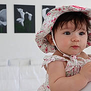 Emma is registered to the contest to win money with this photo: baby, child, floral_hat, floral_dress, indoor, portrait, cute, curious, person, face, skin, hand, earring, artwork, white_background, soft_lighting, infant, closeup, holding, expression