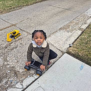 Andrew is registered to the contest to win money with this photo: child, toddler, outdoor, driveway, sidewalk, toy, excavator, motorcycle, scooter, ride_on_toy, standing, kneeling, looking_at_camera, braided_hair, jacket, winter_clothing, residential, brick_house, grass, portrait