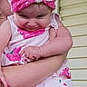 adult_arm, arms, baby, child, closeup, cute, expression, face, floral_pattern, headband, holding, infant, outdoor, person, pink_clothing, portrait, skin, smile, toddler, yellow_wall