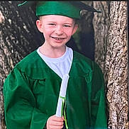 Joshua is registered to the contest to win money with this photo: child, graduate, green_gown, graduation_cap, diploma, tree, outdoor, smiling, portrait, young, celebration, person, happy, nature, childhood, achievement, ceremony, clothing, holding, event