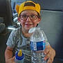 baby_bottle, bottle, boy, bus_seat, casual_clothing, child, daylight, glasses, happy, holding, indoor, portrait, seat, sitting, smiling, transportation, water_bottle, window, yellow_cap, young_child