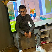 Djilani a rejoint le concours — aidez-le/la à gagner de superbes lots ! boy, child, glasses, sitting, indoor, television, animation, wooden_floor, casual_clothing, sneakers, grey_wall, box, smile, person, living_room, entertainment, screen, furniture, home, young