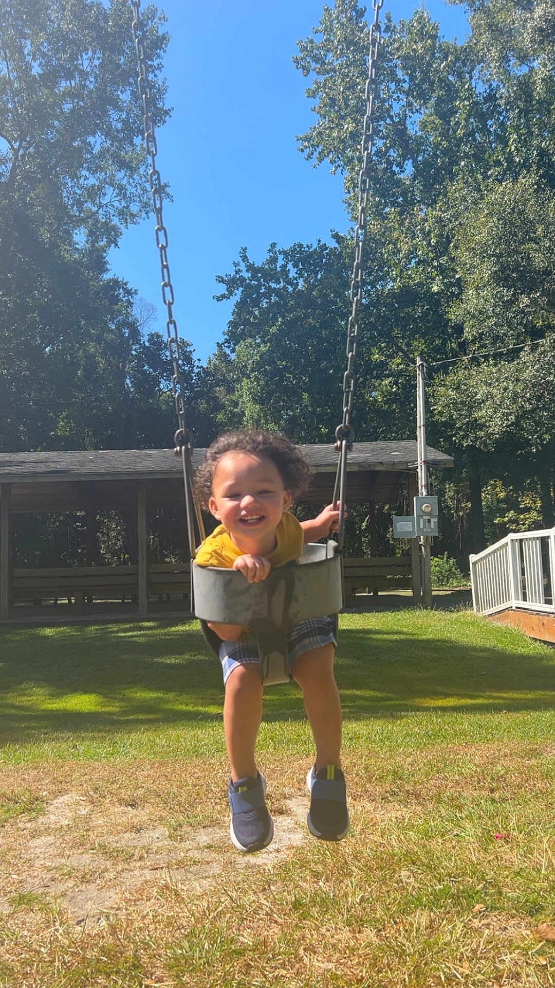 Kyzer is registered to the contest to win money with this photo: child, toddler, swing, playground, outdoor, sunny, grass, trees, park, happy, smiling, casual_clothing, shorts, sneakers, daytime, recreation, fun, motion, joy, nature
