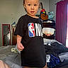 toddler, child, bed, clothing, black_tshirt, nba_logo, container, indoor, mirror, guitar, curtain, furniture, clutter, person, barefoot, room, window, plastic_container, expression, standing