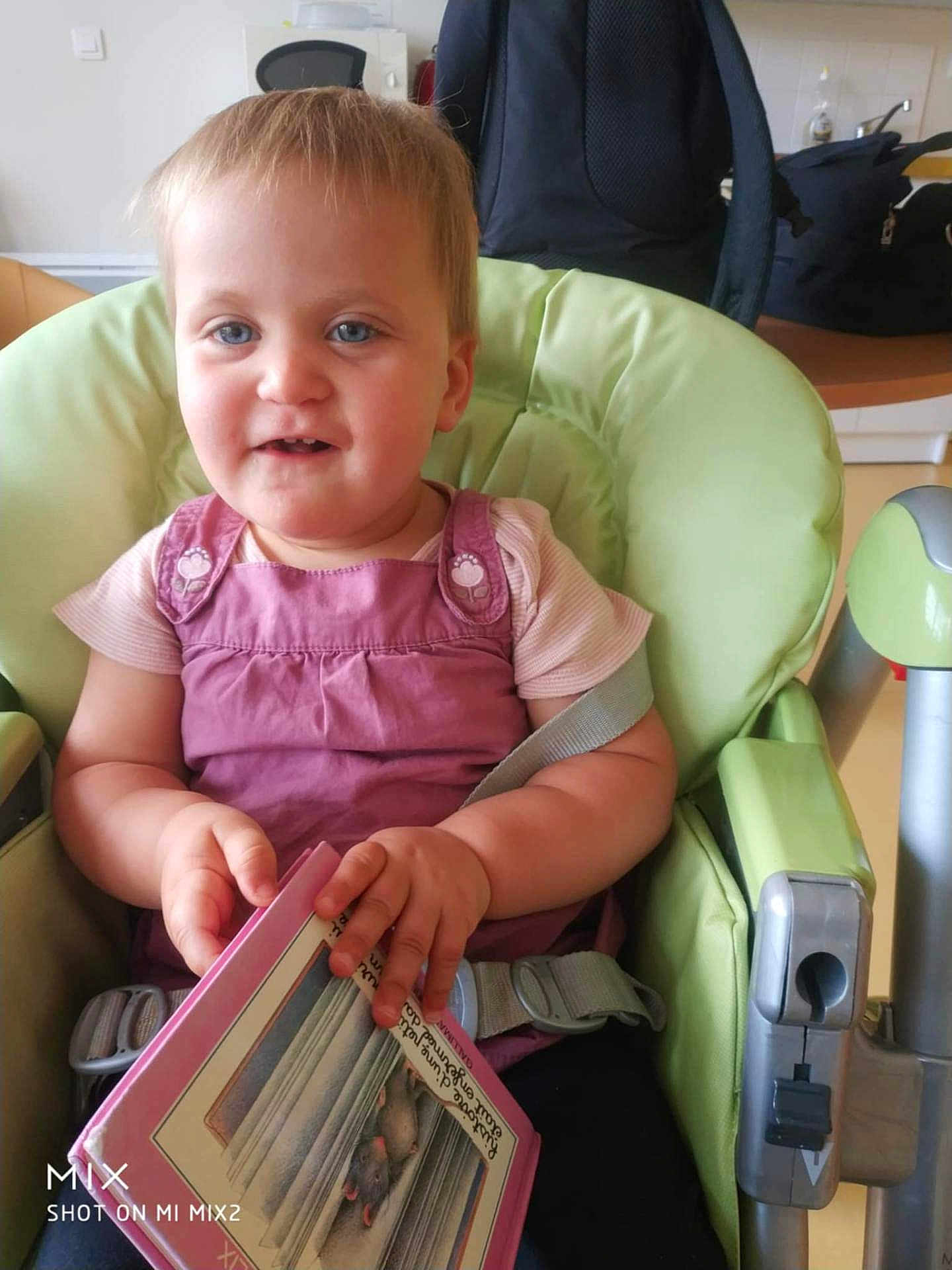 Melyo a rejoint le concours — aidez-le/la à gagner de superbes lots ! toddler, child, high_chair, book, reading, pink_clothing, blue_eyes, indoor, kitchen, baby, seatbelt, furniture, person, portrait, cute, holding, smiling, young_child, domestic, home