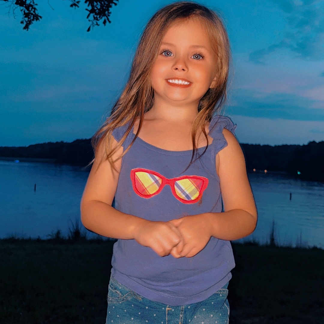 Lillie is registered to the contest to win money with this photo: child, girl, smiling, portrait, outdoor, lakeside, lake, water, evening, sunset, sky, clouds, tree, landscape, standing, shorts, shirt, sunglasses_design, long_hair, nature