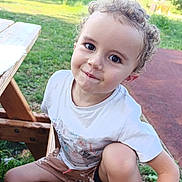 Leo participe au concours pour gagner de l'argent avec cette photo : child, toddler, portrait, outdoor, park, picnic_table, bench, grass, curly_hair, smile, sandals, white_tshirt, shorts, face, big_eyes, playful, candid, nature, happy, sitting