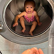 Realynn is registered to the contest to win money with this photo: child, toddler, washing_machine, inside, smiling, happy, playful, hand, metal, appliance, laundry, curious, indoors, clothing, pink_top, barefoot, candid, portrait, baby, sitting