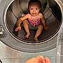 child, toddler, washing_machine, inside, smiling, happy, playful, hand, metal, appliance, laundry, curious, indoors, clothing, pink_top, barefoot, candid, portrait, baby, sitting