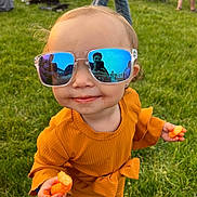 Realynn is registered to the contest to win money with this photo: cheese_puffs, child, cute, event, face, fashion, fun, grass, hand, orange_clothing, outdoor, people, playful, portrait, reflection, smile, snack, sunglasses, sunlight, toddler