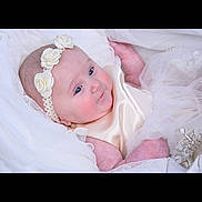 Charlotte is registered to the contest to win money with this photo: baby, infant, headband, flower, white_clothing, silk, soft_fabric, portrait, cute, child, newborn, smile, blue_eyes, skin, lying_down, delicate, tulle, dress, baby_girl, closeup