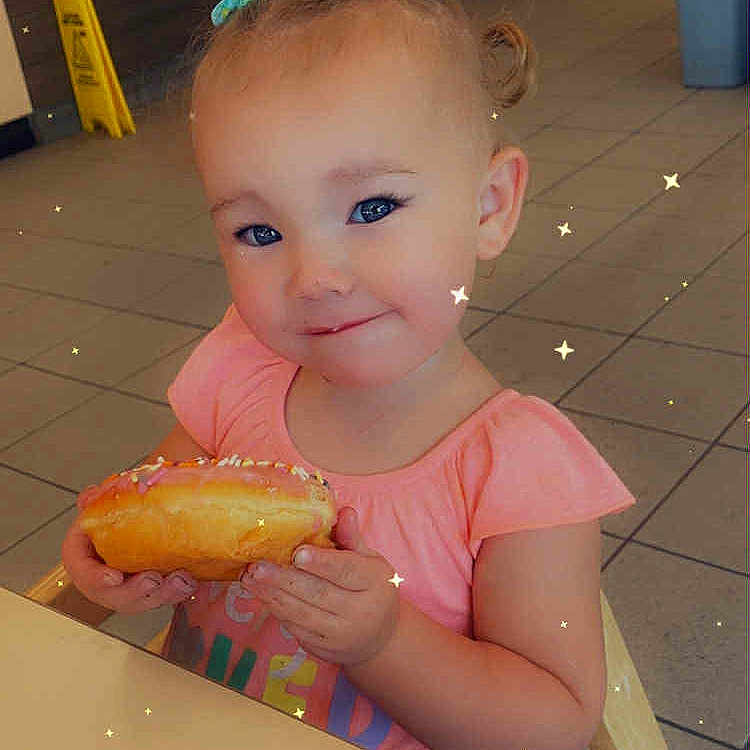 Havanna is registered to the contest to win money with this photo: bakery, child, clothing, cute, donut, face, floor, food, hair_bows, happy, high_chair, indoor, person, pink_shirt, seat, smile, stars, table, tile_floor, toddler