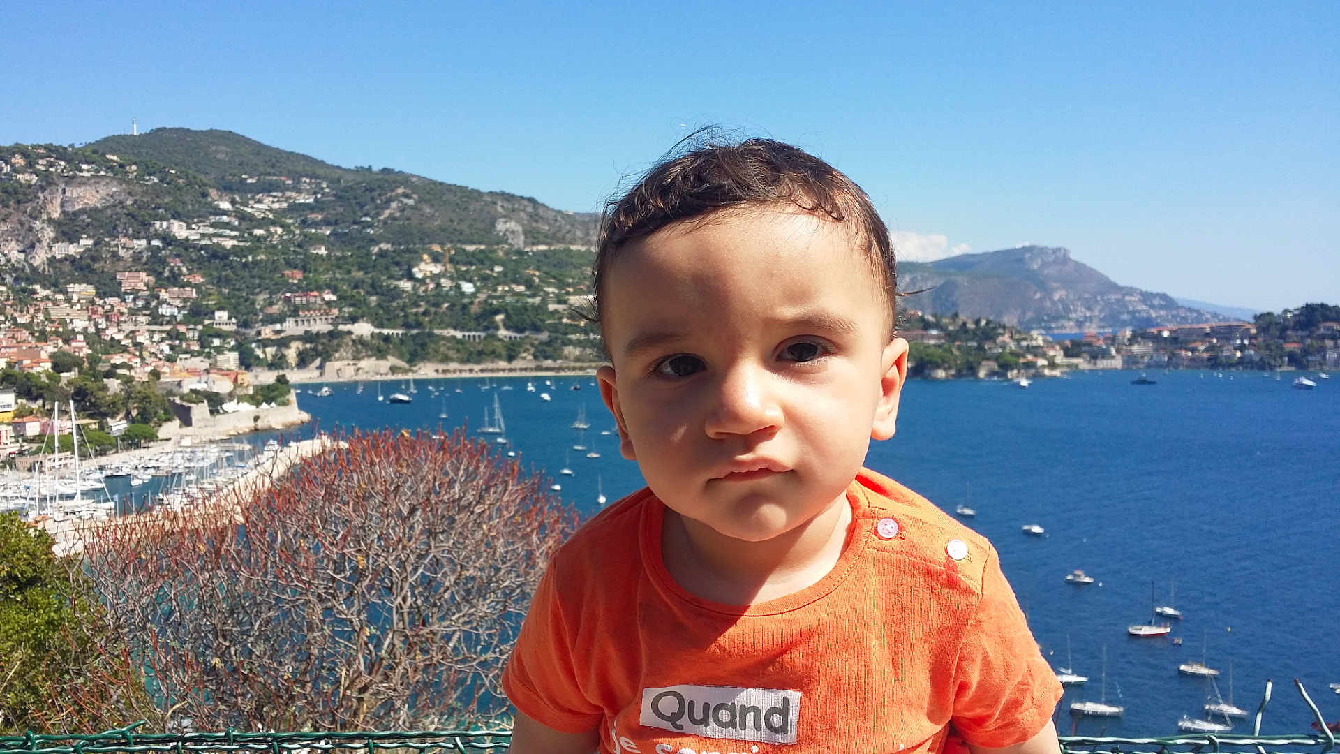 Anis a rejoint le concours — aidez-le/la à gagner de superbes lots ! child, toddler, orange_shirt, face, outdoor, sea, boats, harbor, mountain, sky, daylight, water, nature, person, vacation, travel, landscape, young_child, expression, summer