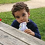 child, curly_hair, outdoor, grass, picnic_table, yogurt_pouch, park, greenery, casual_clothing, curious, innocent, hand, face, nature, daylight, young_child, sitting, playful, relaxed, food