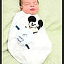 baby, newborn, swaddled, sleeping, blanket, plush_toy, mickey_mouse, face, head, portrait, infant, towel, soft_texture, resting, peaceful, indoors, bedding, photograph, framed_border, watermark