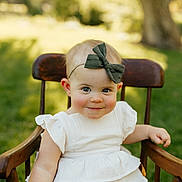 Poppy joined the competition — help win amazing prizes! baby, child, smile, headband, green_bow, white_dress, wooden_chair, outdoor, grass, daylight, portrait, cute, infant, young_child, face, happy, sitting, nature, sunlight, blurry_background
