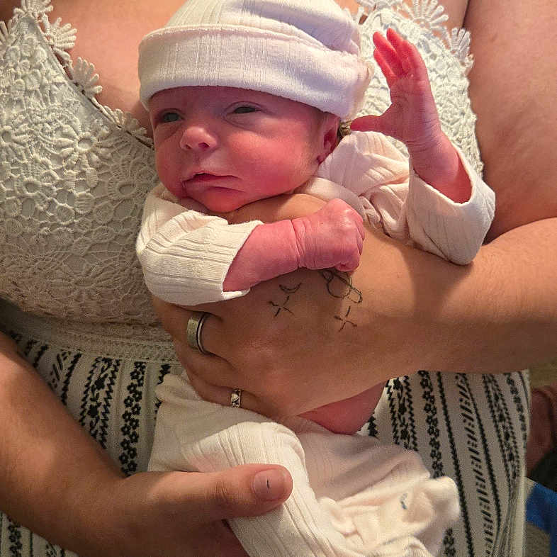 Billie joined the competition — help win amazing prizes! adult, baby, newborn, holding, hat, tattoo, necklace, white_clothing, striped_skirt, indoor, person, portrait, hands, skin, face, bed, blanket, casual, warm_lighting, closeup