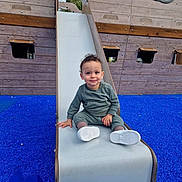 Jad participe au concours pour gagner de l'argent avec cette photo : toddler, child, slide, playground, blue_surface, wooden_structure, pirate_ship, smile, curly_hair, green_outfit, white_shoes, outdoor, play, fun, happy, person, recreation, park, daytime, young