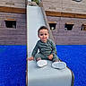 toddler, child, slide, playground, blue_surface, wooden_structure, pirate_ship, smile, curly_hair, green_outfit, white_shoes, outdoor, play, fun, happy, person, recreation, park, daytime, young