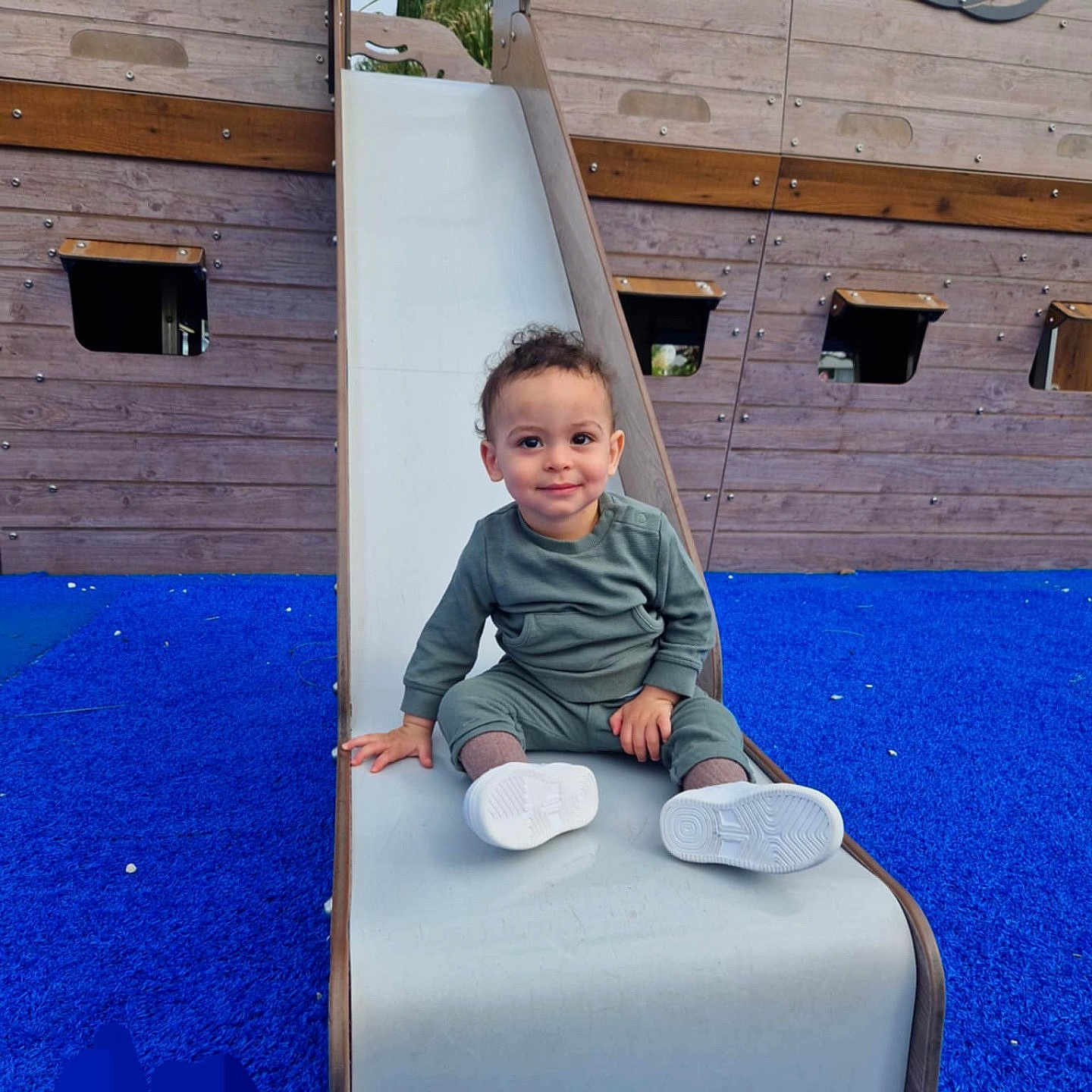 Jad participe au concours pour gagner de l'argent avec cette photo : toddler, child, slide, playground, blue_surface, wooden_structure, pirate_ship, smile, curly_hair, green_outfit, white_shoes, outdoor, play, fun, happy, person, recreation, park, daytime, young