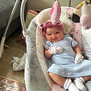 Lily is registered to the contest to win money with this photo: baby, infant, smile, mittens, headband, baby_rocker, soft_toys, striped_dress, white_socks, indoor, floor, carpet, cushion, baby_accessories, child, cute, relaxing, portrait, happy, cozy