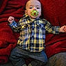 baby, pacifier, plaid_shirt, yellow, blue, gray_pants, red_blanket, infant, child, lying_down, soft_texture, cozy, cute, indoors, portrait, young_child, casual_clothing, relaxed, small_hands, socks