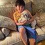 boy, dog, couch, pillow, smiling, indoor, child, pet, casual_clothing, happy, fur, shorts, orange_shirt, relaxed, seated, home, living_room, companion, cozy, friendship