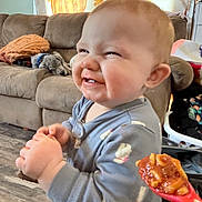 Octavia is registered to the contest to win money with this photo: baby, toddler, smiling, messy_face, food, spoon, sauce, pajamas, living_room, couch, blanket, curtain, pumpkin, decor, laundry_basket, standing, hands, tooth, floor, indoor