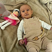 Julia participe au concours pour gagner de l'argent avec cette photo : baby, blanket, doll, pacifier, overalls, infant, toy, child, soft_texture, beige, pink, smiling, lying_down, indoors, cute, clothing, face, person, young_child, cozy