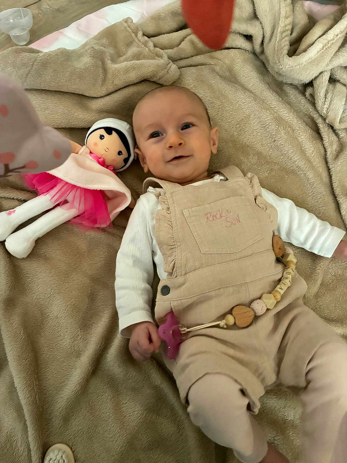 Julia participe au concours pour gagner de l'argent avec cette photo : baby, blanket, doll, pacifier, overalls, infant, toy, child, soft_texture, beige, pink, smiling, lying_down, indoors, cute, clothing, face, person, young_child, cozy