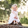 child, girl, dress, outdoor, nature, tree_stump, smile, sunlight, forest, blurry_background, happy, cute, portrait, sitting, young, shoes, pigtails, playful, daylight, grass