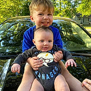 Ezekiel joined the competition — help win amazing prizes! baby, child, sibling, smiling, outdoor, car, hood, greenery, trees, sunlight, happy, holding, cute, portrait, blue_shirt, onesie, hands, face, nature, family