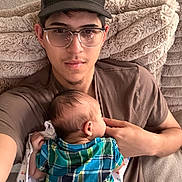 Kai Lovato is registered to the contest to win money with this photo: baby, infant, young_man, father, parent, cuddling, portrait, selfie, glasses, baseball_cap, plaid_shirt, blanket, couch, indoor, sitting, family, 抱抱, cozy, caregiving, photograph