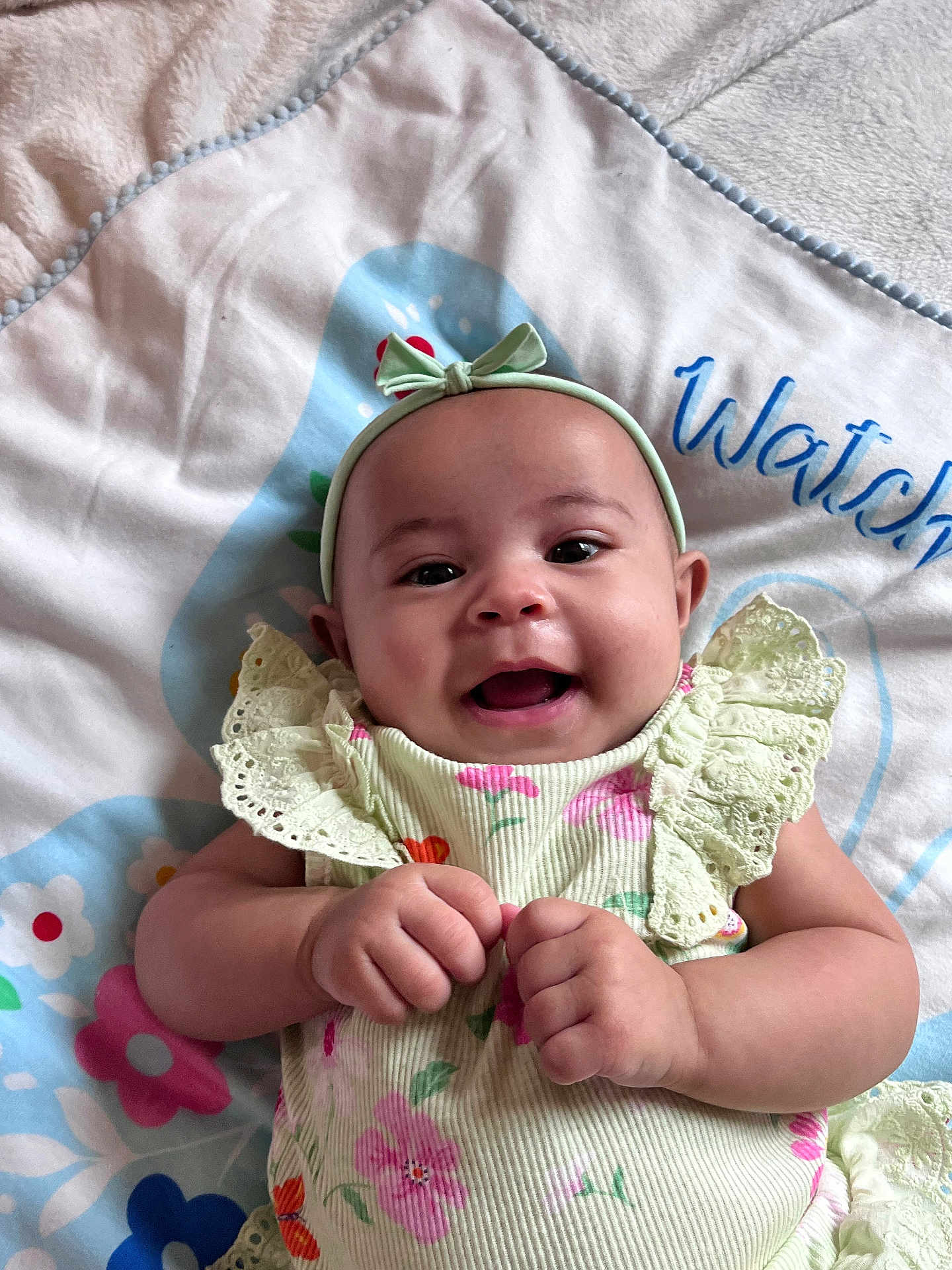 Ivy is registered to the contest to win money with this photo: baby, smiling, infant, headband, bow, floral_dress, hands, blanket, pattern, colorful, happy, cute, lying_down, young_child, soft_texture, indoors, portrait, face, skin, child