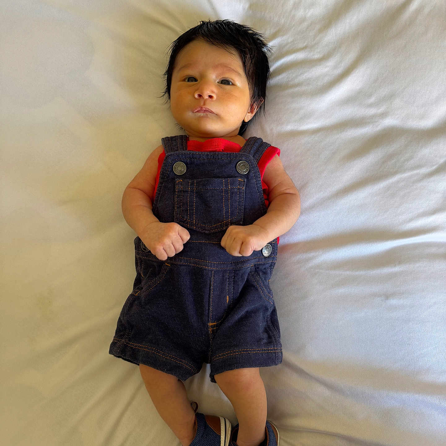 Gabriel is registered to the contest to win money with this photo: baby, infant, denim_overalls, red_shirt, blue_shoes, lying_down, blanket, indoor, cute, serious_expression, clothing, footwear, small_hands, portrait, child, young, soft_lighting, casual_wear, fashion, newborn