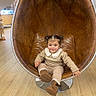 boots, chair, child, collar, cozy, egg_chair, face, girl, hand, indoor, leather, outfit, pigtails, playful, retail_display, sitting, smile, store, toddler, wooden_floor