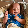 baby, child, infant, smiling, happy, portrait, sitting, bouncer, baby_seat, blue_clothing, patterned_clothing, raised_hand, indoor, home, cushion, safety_harness, furniture, warm_lighting, close_up, photography
