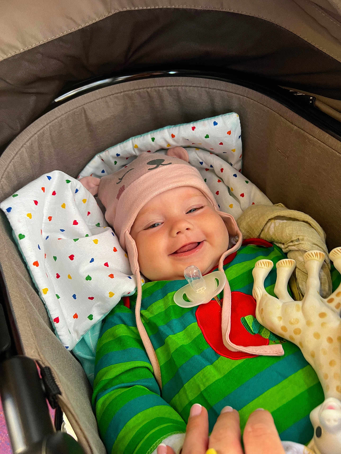 Ivy is registered to the contest to win money with this photo: baby, smiling, hat, stroller, blanket, toy, giraffe, green_stripes, red_apple, cute, happy, child, infant, pacifier, pink_hat, cozy, colorful, heart_pattern, hand, lying_down