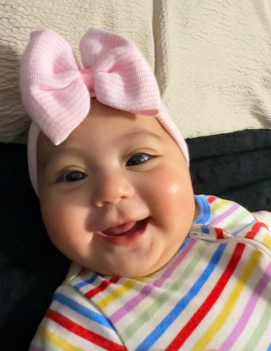 Meghan is registered to the contest to win money with this photo: baby, infant, child, bow_headband, pink_bow, smile, chubby_cheeks, onesie, striped_clothing, close_up, portrait, indoor, blanket, bed, happy, eyes, mouth, skin, cute, head