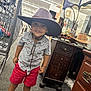 child, boy, cowboy_hat, oversized_hat, smile, thrift_store, shopping_cart, aisle, antique_furniture, mirror, sandals, red_shorts, striped_shirt, carpet_floor, playful, portrait, candid, retail, hat_chin_strap, footwear