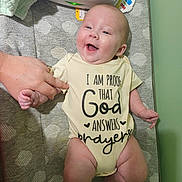 Hazel is registered to the contest to win money with this photo: baby, infant, smiling, onesie, hand, changing_pad, pattern, happy, person, indoor, fabric, text_on_clothing, skin, adult_hand, blanket, baby_care, cute, child, clothing, lying_down