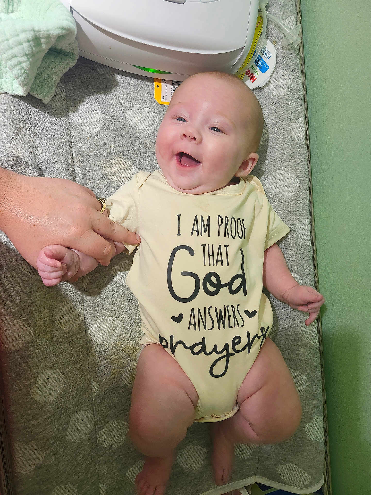 Hazel is registered to the contest to win money with this photo: baby, infant, smiling, onesie, hand, changing_pad, pattern, happy, person, indoor, fabric, text_on_clothing, skin, adult_hand, blanket, baby_care, cute, child, clothing, lying_down