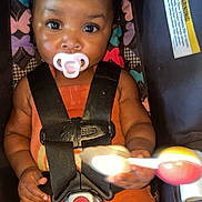 Milianah is registered to the contest to win money with this photo: baby, child, pacifier, car_seat, safety_harness, rattle_toy, orange_clothing, braided_hair, patterned_background, indoor, cute, infant, playful, colorful, closeup, portrait, smiling_eyes, seated, young_child, secure