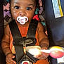 baby, child, pacifier, car_seat, safety_harness, rattle_toy, orange_clothing, braided_hair, patterned_background, indoor, cute, infant, playful, colorful, closeup, portrait, smiling_eyes, seated, young_child, secure